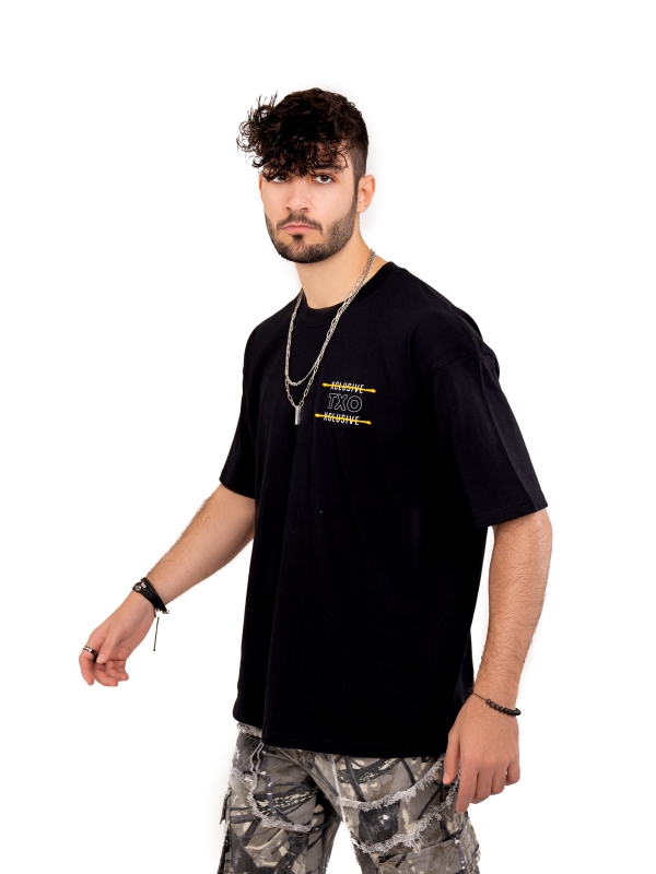 Man With Curly Hair Wearing A Black Graphic T Shirt And Camouflage Trousers Against A Plain White Background.